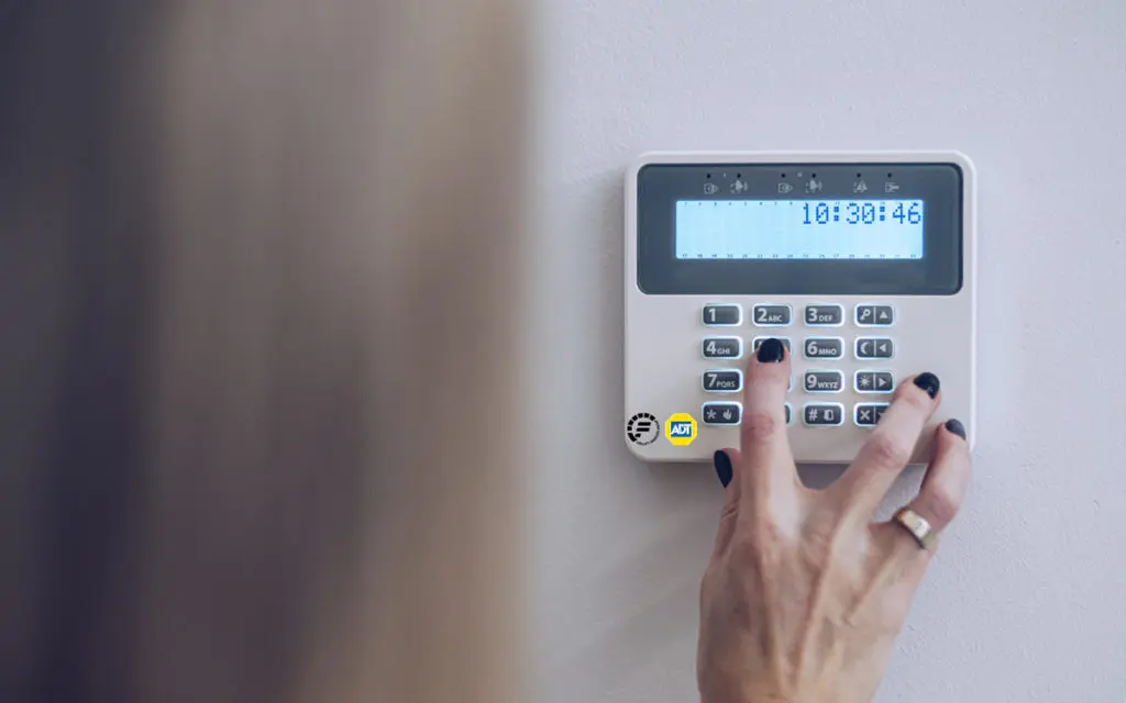 Hand pressing buttons on a wall-mounted security keypad (ADT) with a blue LCD display showing time.