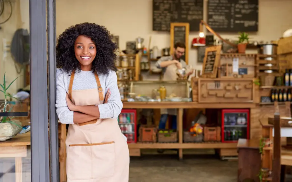 Smiling woman with curly hair in a beige apron stands at the doorway of a cozy cafe, with a wooden counter and shelves behind her.