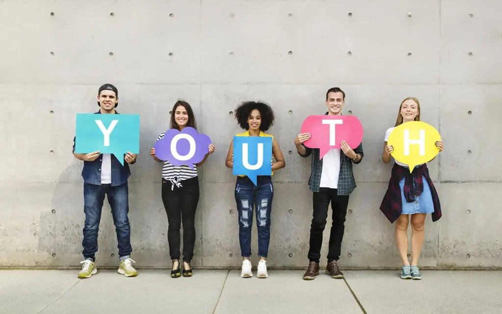Five young adults stand against a concrete wall, each holding a colored speech bubble with letters spelling 'YOUTH'.