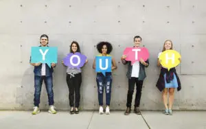 Five young adults stand against a concrete wall, each holding a colored speech bubble with letters spelling 'YOUTH'.