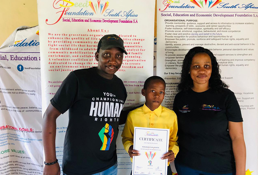 Power-of-mentorship-4 - Fidelity ADT - South African Home & Commercial Security Three people pose for a photo at a community foundation event; a child in yellow holds a certificate between two adults with banners in the background.