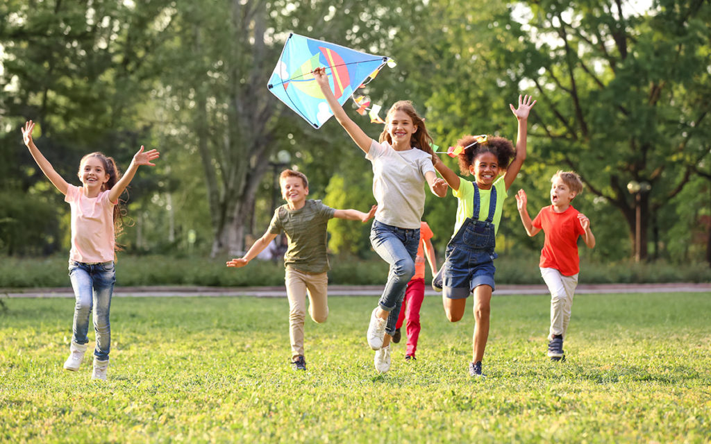 Group of children running and laughing in a sunny park, flying a colorful diamond-shaped kite.