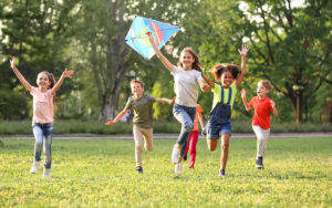 Group of children running and laughing in a sunny park, flying a colorful diamond-shaped kite.