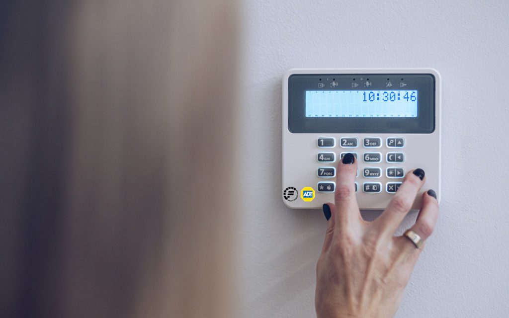 Hand pressing buttons on a wall-mounted security keypad (ADT) with a blue LCD display showing time.