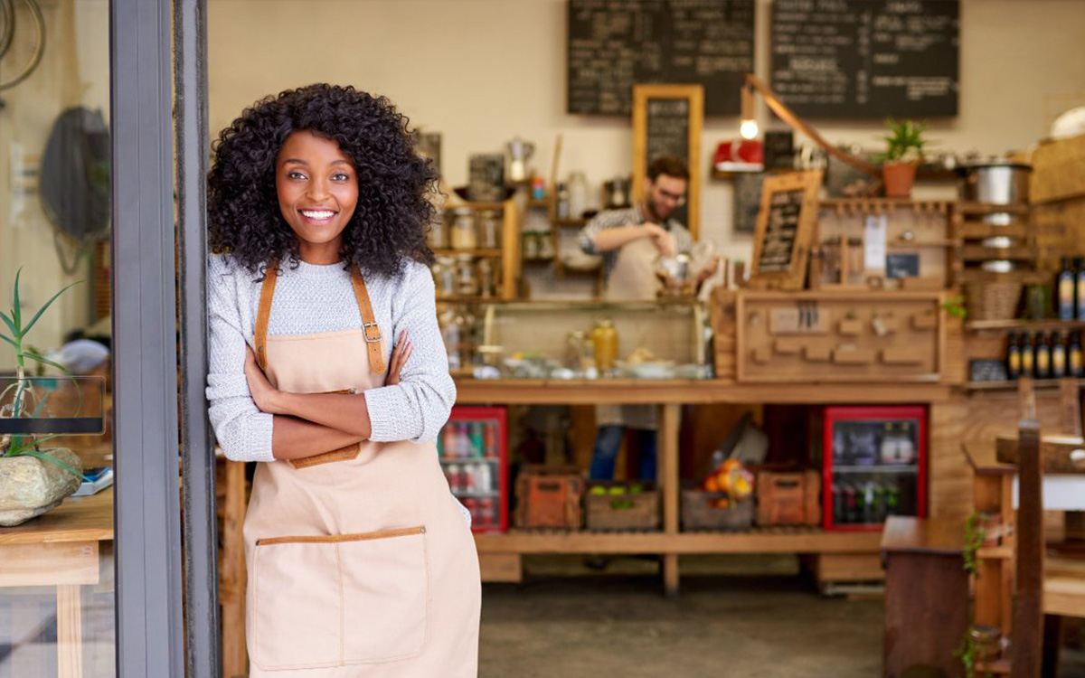 business-safety - Fidelity ADT - South African Home & Commercial Security - Fidelity ADT - South African Home & Commercial Security Smiling woman with curly hair in a beige apron stands at the doorway of a cozy cafe, with a wooden counter and shelves behind her.