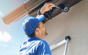 Blue-shirted worker installs a security camera under the eaves of a house, with a ladder nearby in the sunlight.