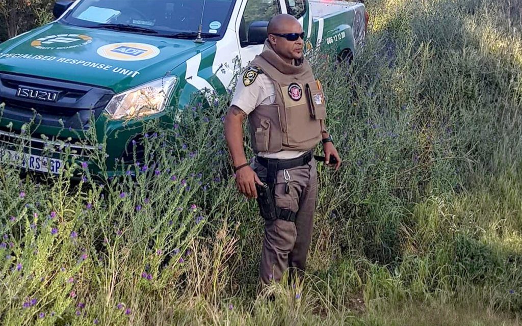 rural-safety2 - Fidelity ADT - South African Home & Commercial Security Uniformed security officer in beige tactical vest and sunglasses stands in tall grass beside a green and white patrol vehicle marked 'Specialised Response Unit.'