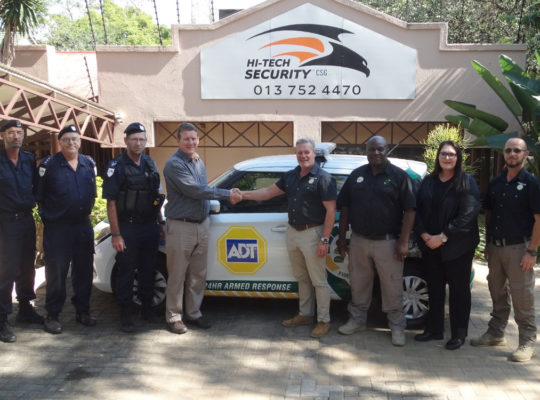 Group of security guards posing in front of a Hi-Tech Security sign and an ADT patrol car; two men shake hands in the center.