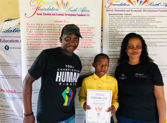 Three people pose for a photo at a community foundation event; a child in yellow holds a certificate between two adults with banners in the background.