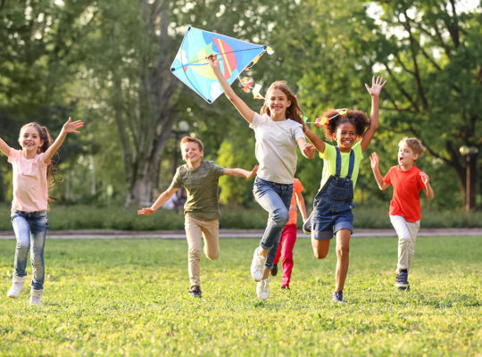 Group of children running and laughing in a sunny park, flying a colorful diamond-shaped kite.