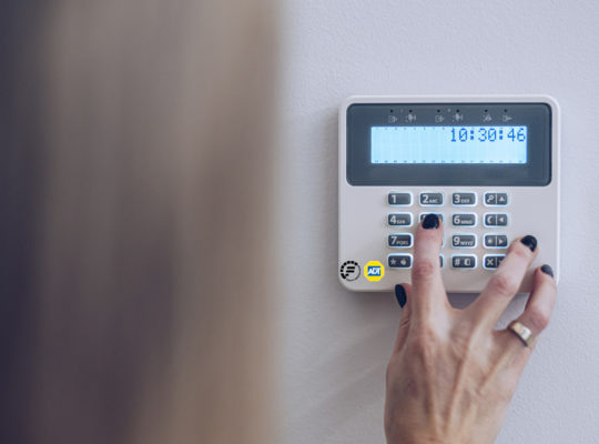 Hand pressing buttons on a wall-mounted security keypad (ADT) with a blue LCD display showing time.