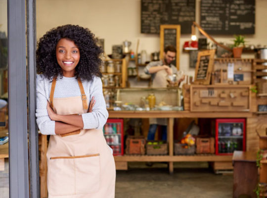 Smiling woman with curly hair in a beige apron stands at the doorway of a cozy cafe, with a wooden counter and shelves behind her.