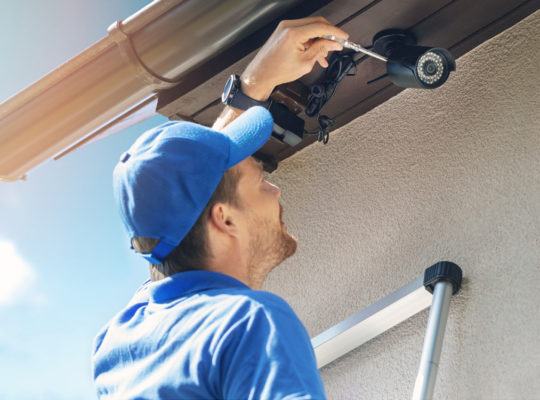 Blue-shirted worker installs a security camera under the eaves of a house, with a ladder nearby in the sunlight.