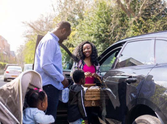 The young family gets out of the family car ready for a day out in a public park. It's a sunny day and they are all happy wearing casual clothing.