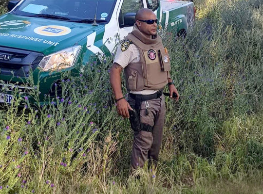 Uniformed security officer in beige tactical vest and sunglasses stands in tall grass beside a green and white patrol vehicle marked 'Specialised Response Unit.'