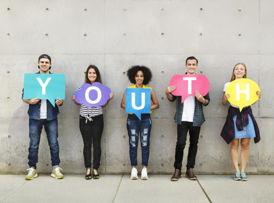 Five young adults stand against a concrete wall, each holding a colored speech bubble with letters spelling 'YOUTH'.